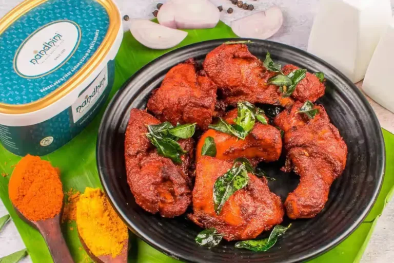 A plate of Andhra-style chicken fry coated in red Guntur-chilli masala with curry leaves, beside a Nandhini Deluxe branded tub and spoons of red and turmeric powder.