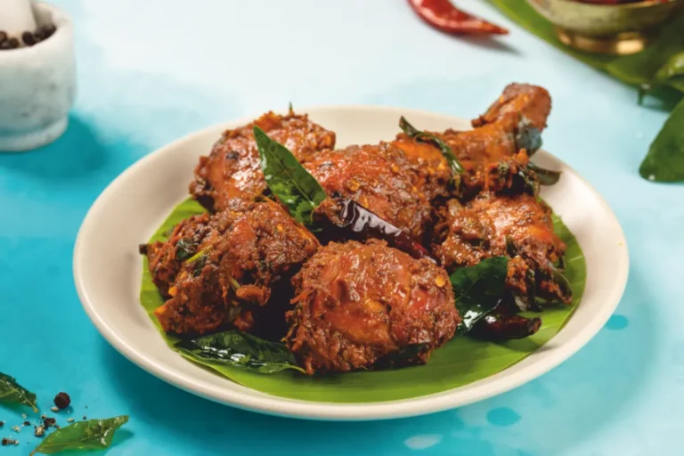 Andhra-style chicken pieces coated in dark red masala and curry leaves, served on a banana leaf over a white plate.