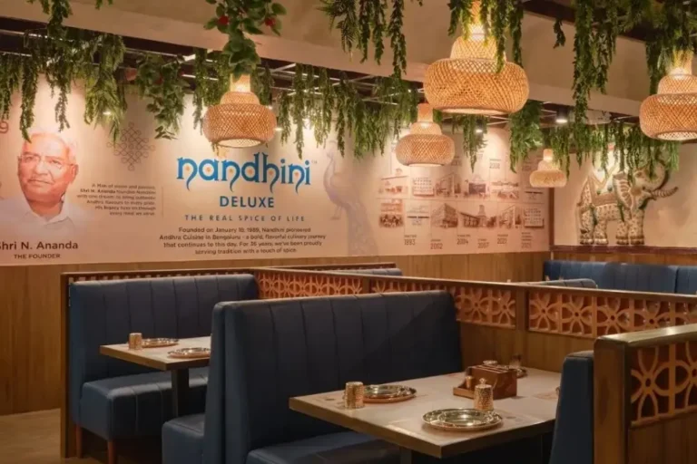 Interior of a modern Nandhini Deluxe outlet with blue booth seating, woven pendant lights, and a feature wall showing the founder's portrait and brand timeline.
